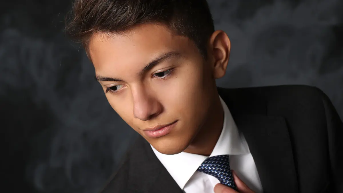 Tight close-up of a senior young man in a dark suit, white shirt and navy patterned tie, hand adjusting the tie, eyes cast down and to the left