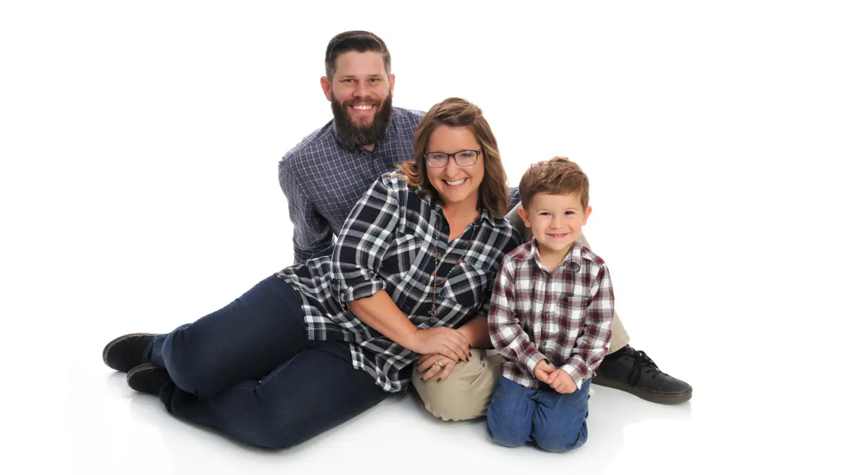 Family of three (man, woman, small boy) in matching plaid flannel shirts and jeans, seated/lying close together on a white seamless backdrop