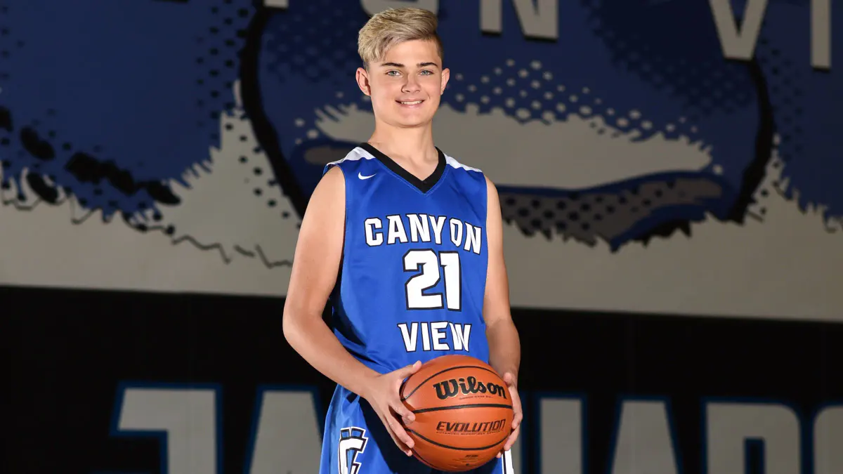 Teen basketball player in a blue 'CANYON VIEW 21' uniform holding a Wilson ball, standing in a gym in front of a large blue Jaguars wall mural