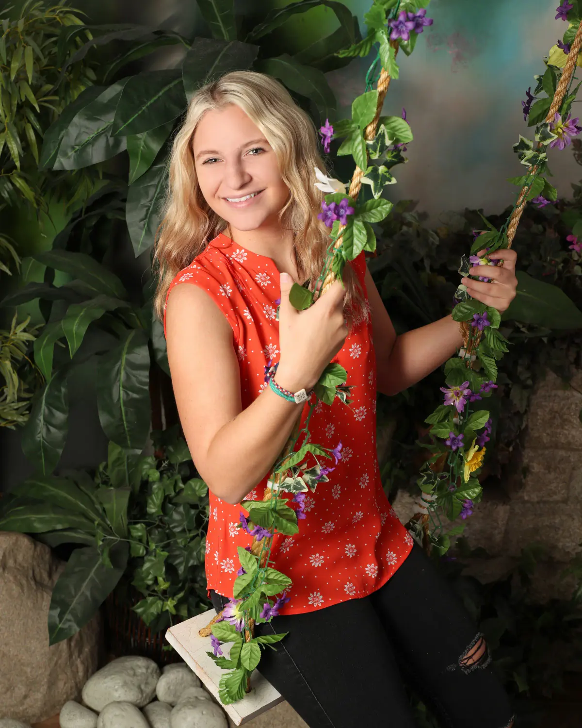 Blonde young woman in red-orange floral sleeveless top and ripped jeans sitting on an indoor swing wrapped in faux purple-flower vines