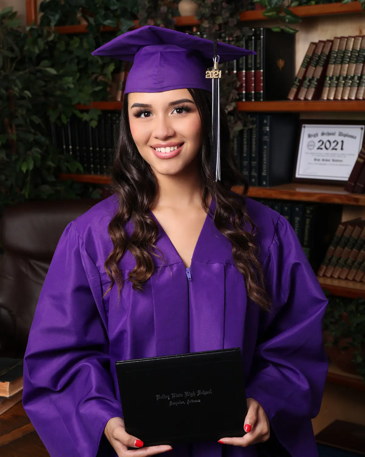 Smiling young woman with long dark wavy hair in purple cap and gown with '2021' year charm on tassel, holding a black diploma cover