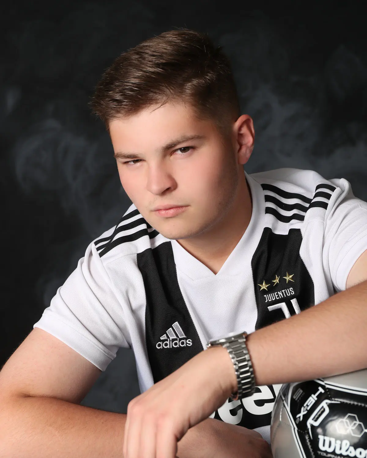 Young man in white/black striped Juventus Adidas soccer jersey and wristwatch, arms crossed resting on a soccer ball, serious expression