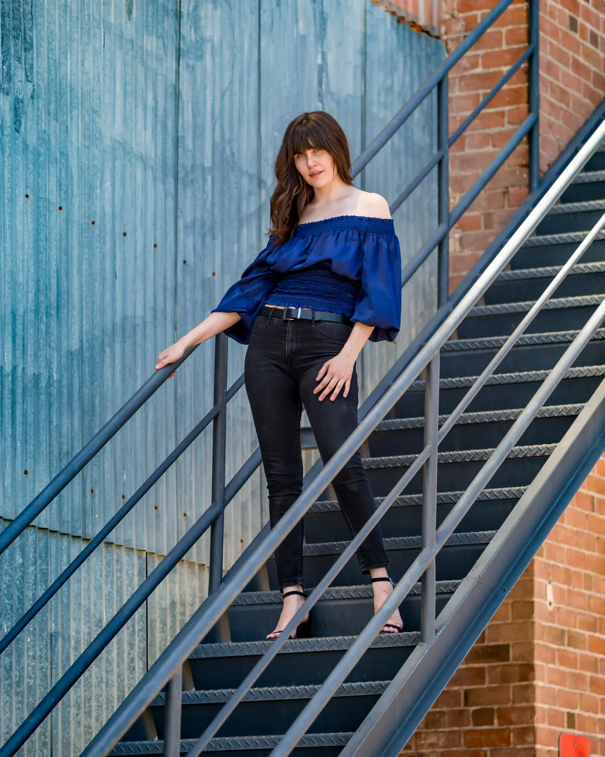 Young woman with long dark hair in royal-blue off-shoulder ruffle top, black jeans and heeled sandals