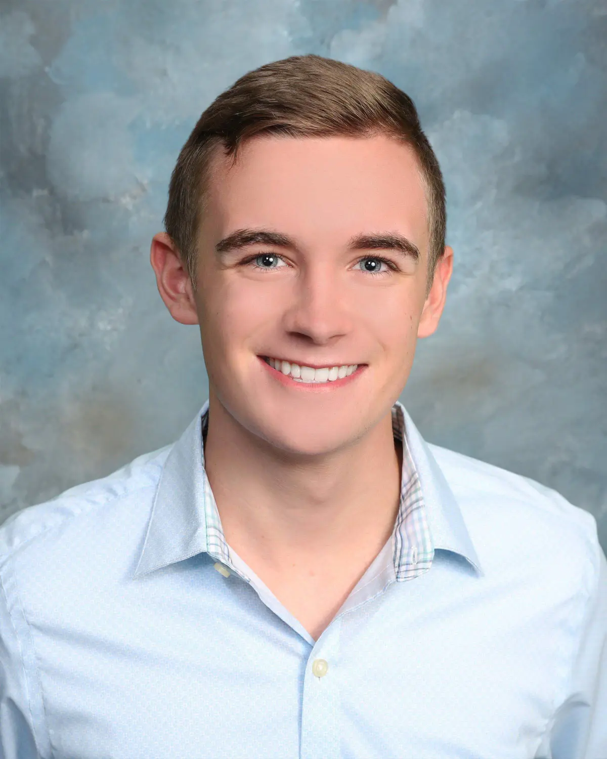 Young man with short brown hair in light blue button-down shirt, big smile, classic head-and-shoulders on blue mottled backdrop; yearbook-style