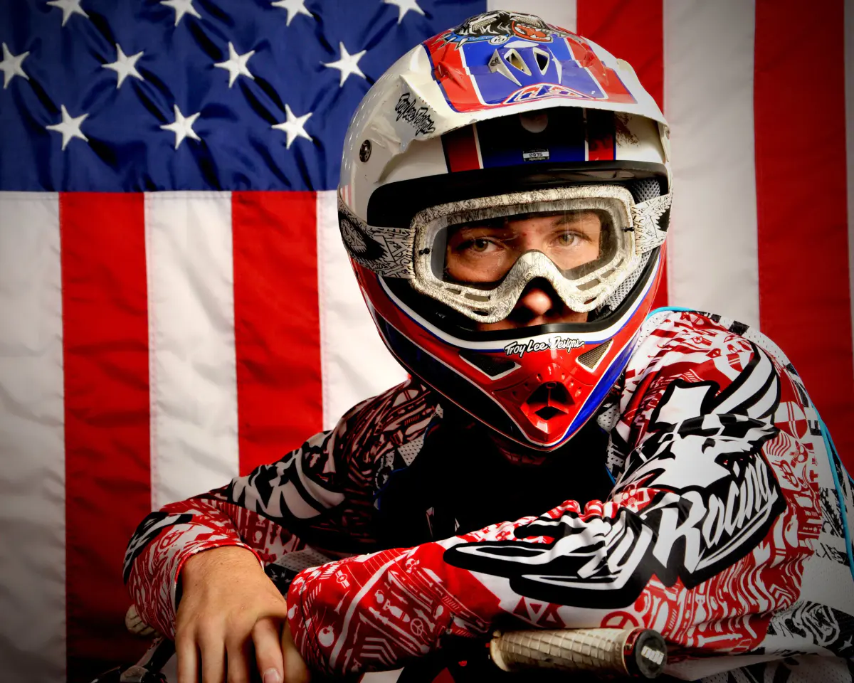 Motocross rider in red/white/blue Troy Lee Designs helmet and goggles and Fly Racing jersey, leaning on handlebars, eyes visible through goggles