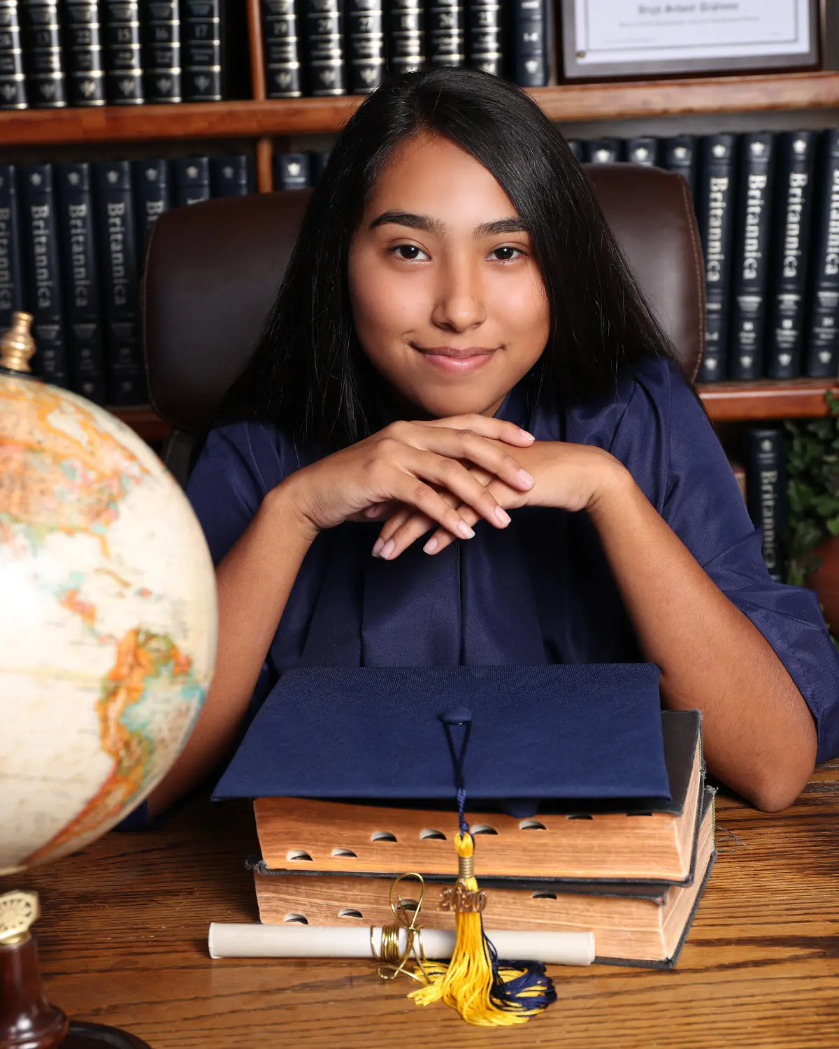 Young woman in navy graduation gown sitting at a wooden desk, chin on folded hands