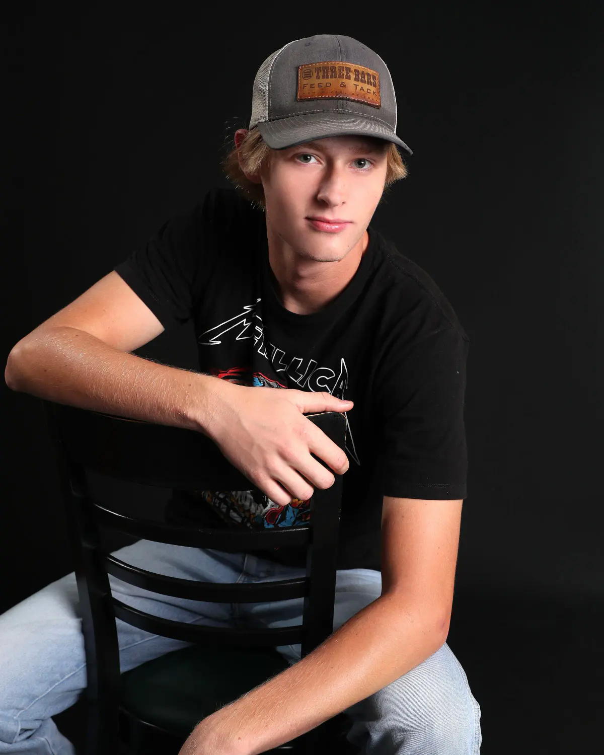Senior guy in a trucker cap, black graphic tee and jeans, sitting backwards on a black wooden chair with arms folded over the backrest