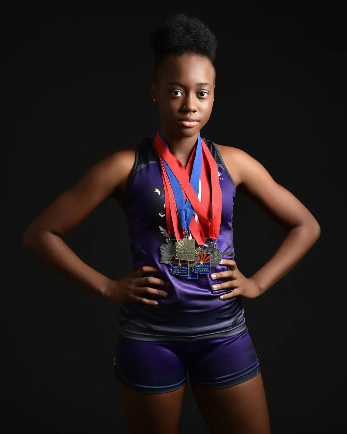 Track athlete in a purple singlet and shorts, hands on hips, steady direct gaze, several medals on red/blue ribbons around her neck