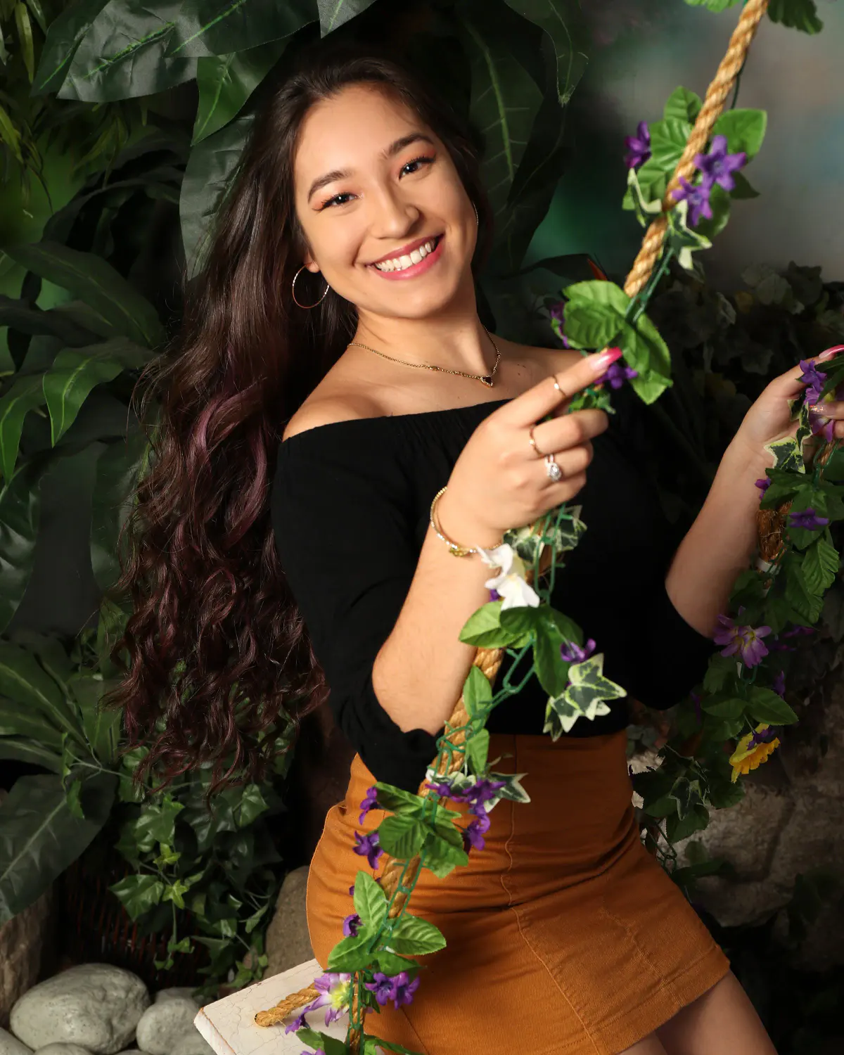 Smiling young woman with long dark hair, black off-shoulder top and rust skirt, on a rope swing wrapped in purple flowers and ivy