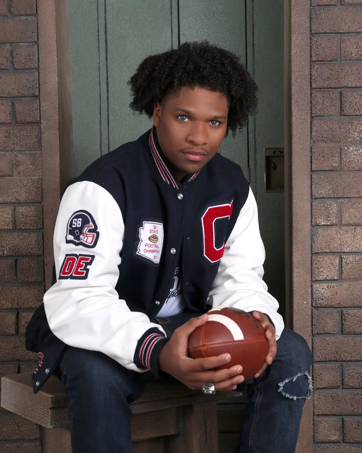 Senior in a navy/white varsity letter jacket with patches (red C, 'DE', helmet #56, '5A State Football Champions 2017')