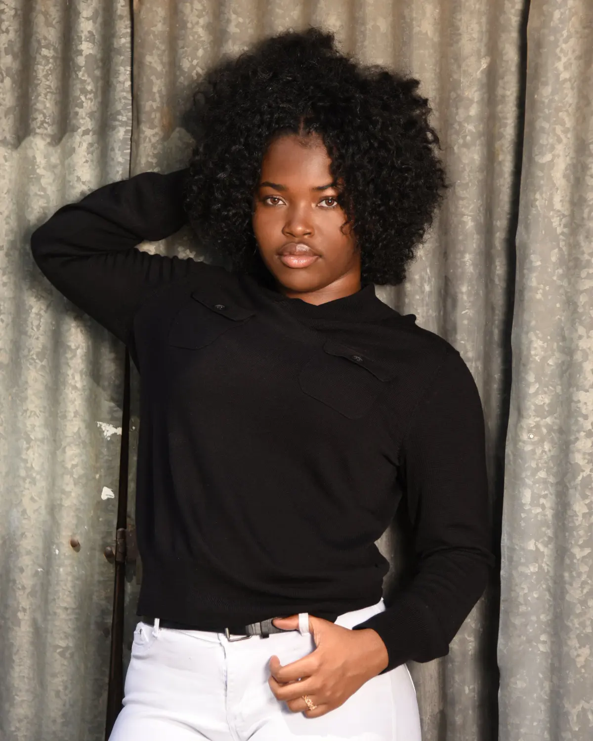 Young woman with natural curly hair in a black sweater with button flap pockets and white jeans, one arm up behind her head, serious direct gaze