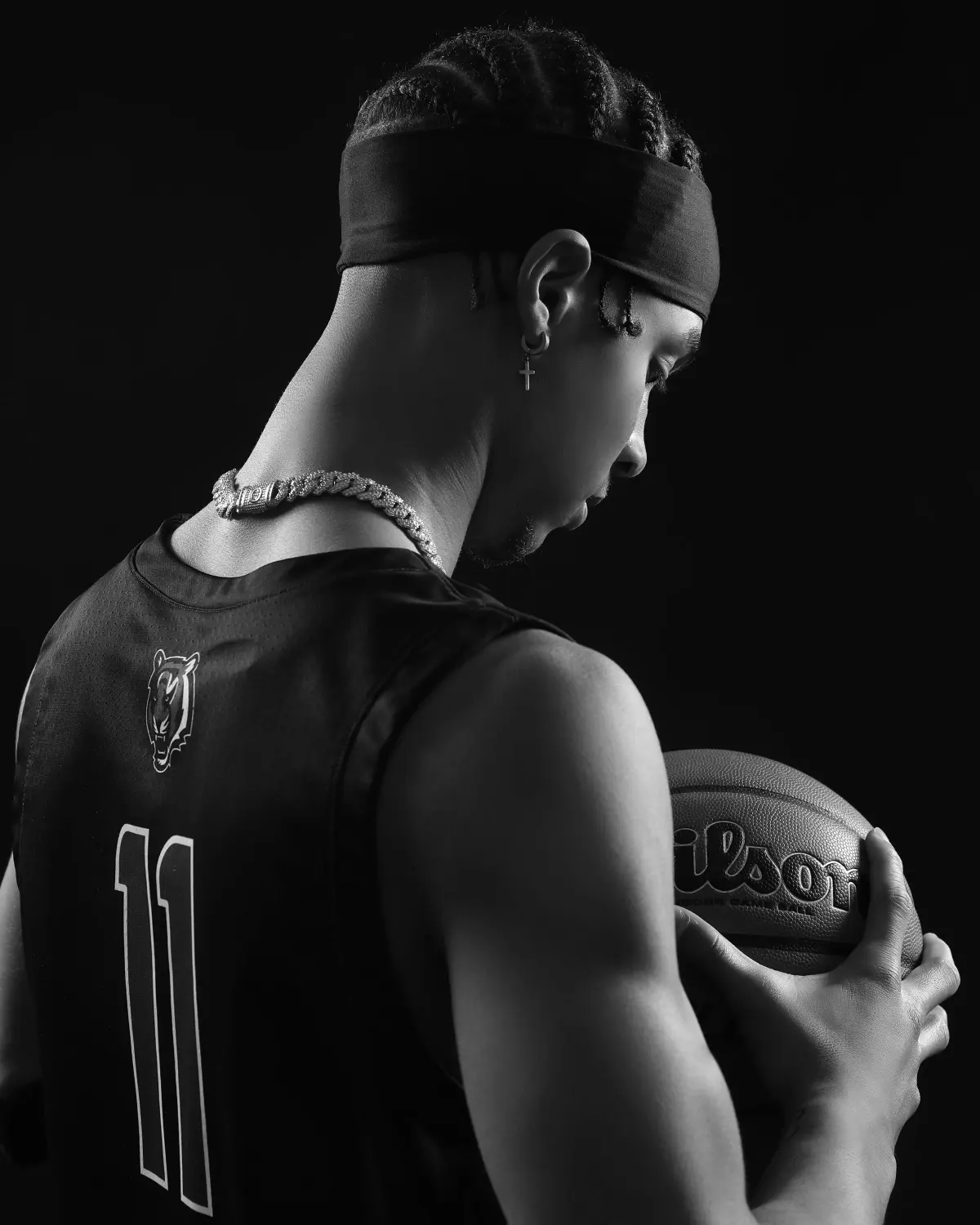 Black-and-white low-key portrait of a basketball player seen from behind at three-quarter angle, head bowed, black headband, braids, cross earring