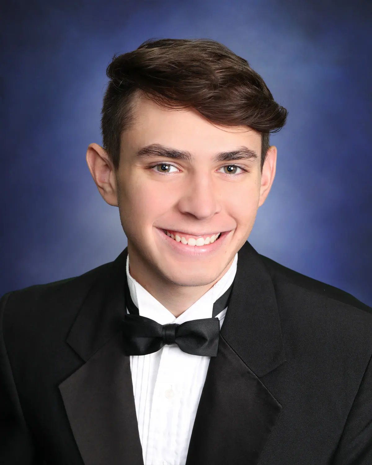 Head-and-shoulders senior portrait of a smiling young man in a black tuxedo with bow tie, against a mottled blue painted backdrop