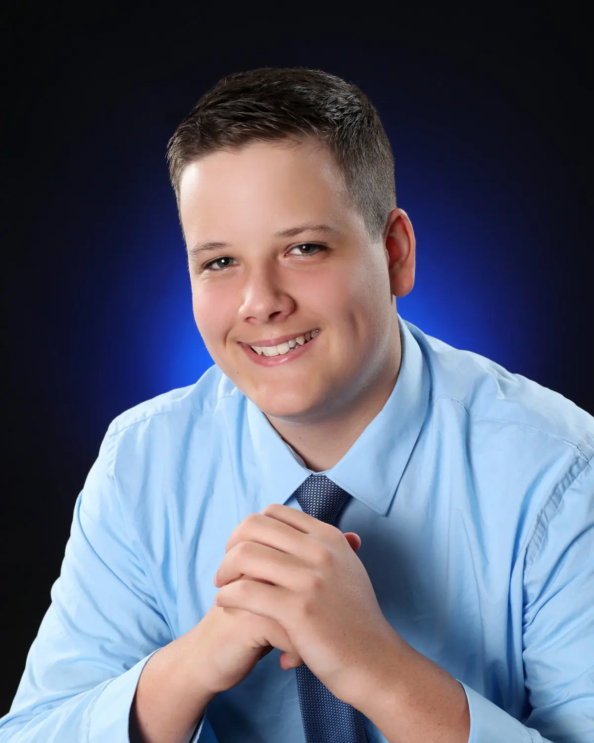 Senior young man in a light blue shirt and patterned tie, hands clasped at chin height and leaning forward, smiling