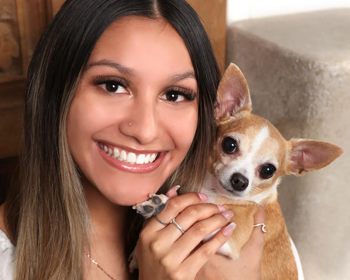 Close-up of a smiling young woman holding a small chihuahua next to her face, against a warm cream/beige background