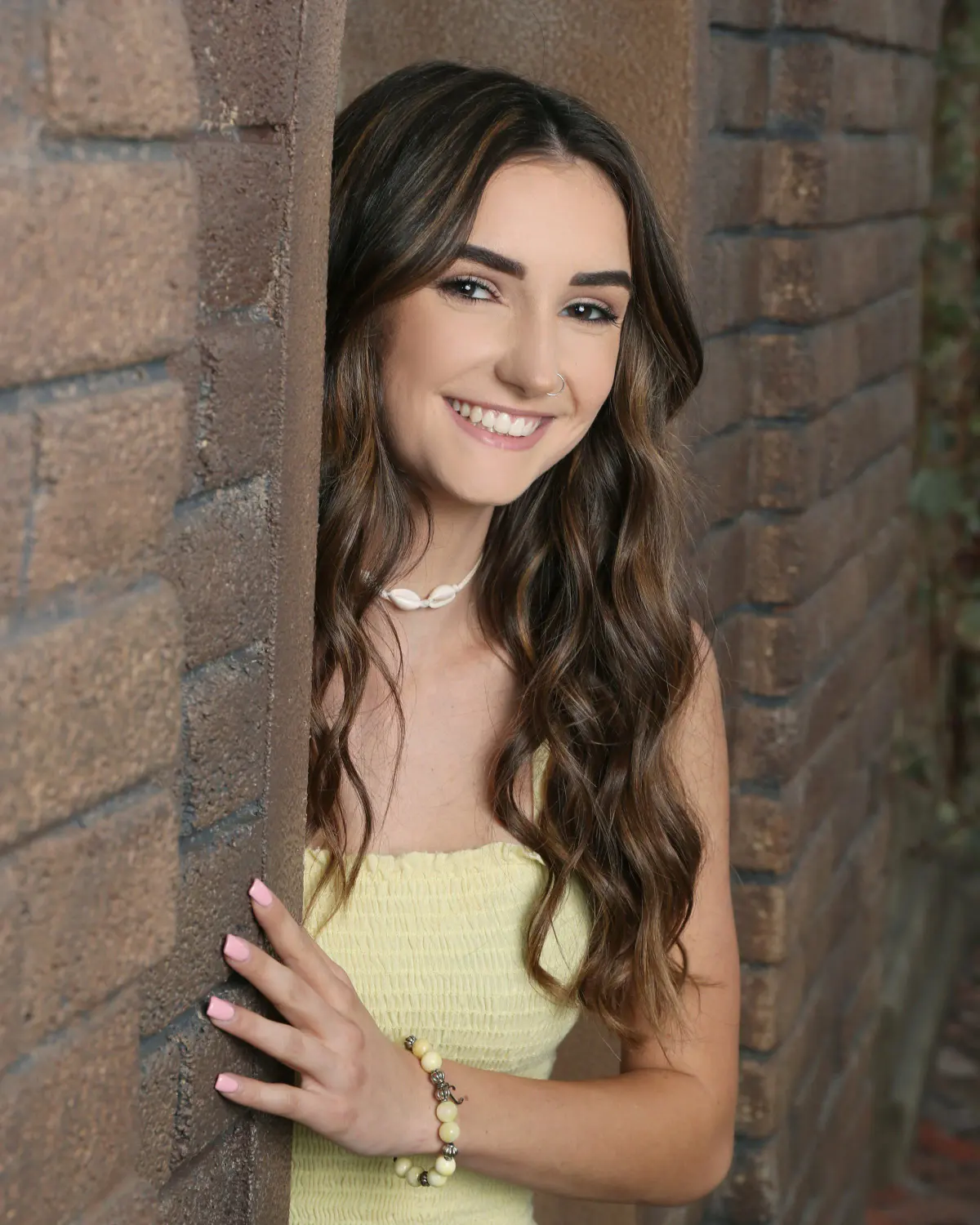 Smiling young woman with long wavy hair in a pale yellow top, leaning against a brick column/doorway on what appears to be an indoor set
