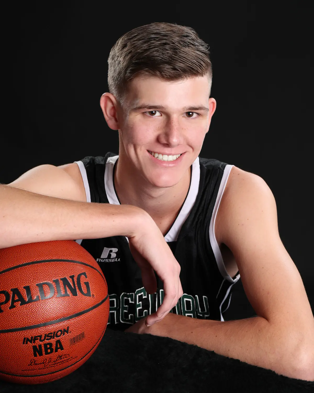 Smiling senior young man in a black basketball jersey leaning on a basketball (Spalding/NBA visible) on a black backdrop