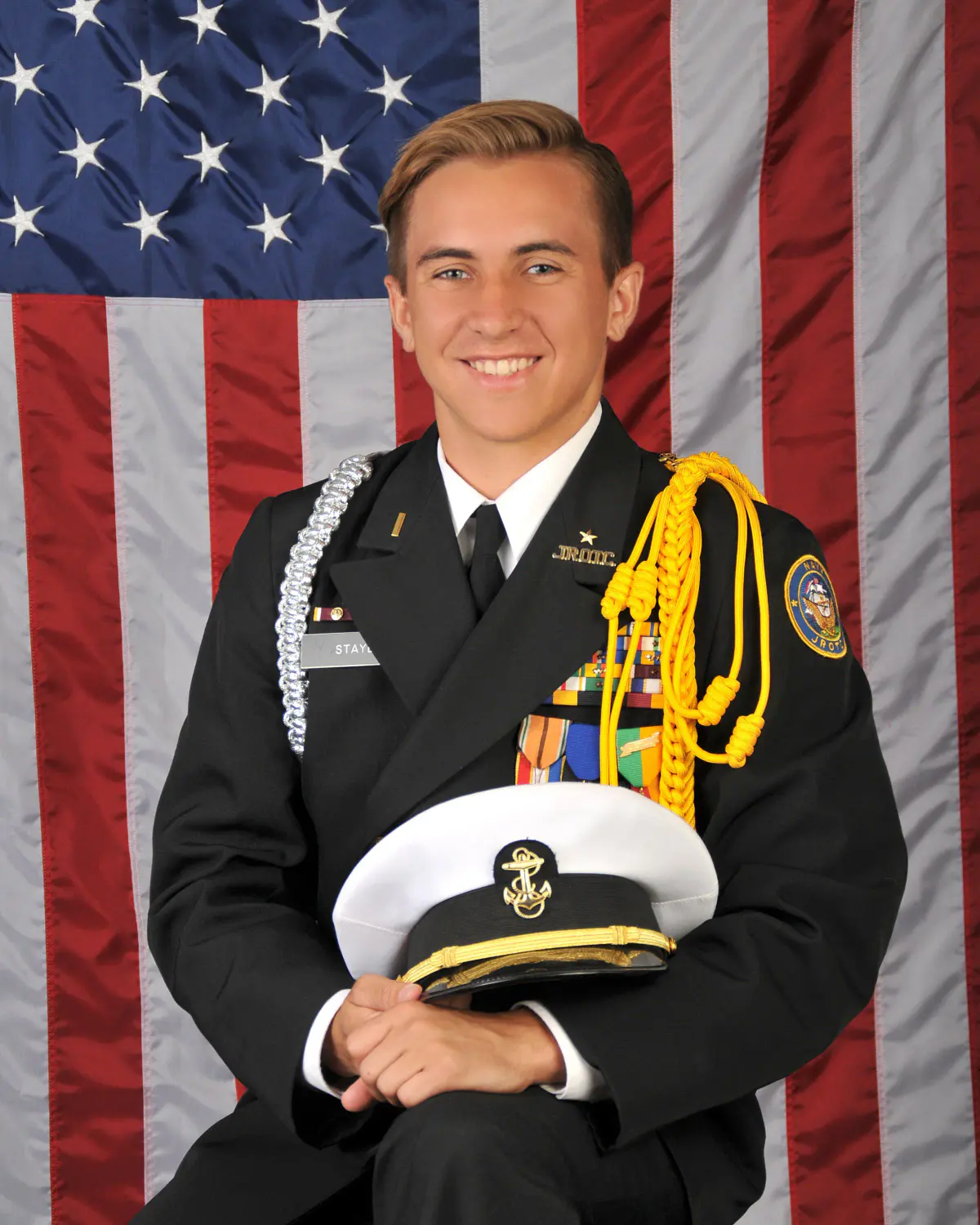Senior young man in navy-style dress uniform with aiguillette cords, ribbons and medals, holding a white combination cover (hat) in lap