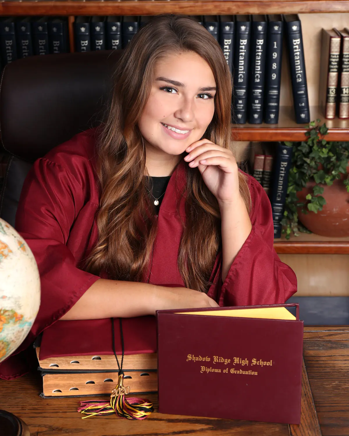 Senior girl with long wavy hair in a maroon graduation gown draped open, leaning on a desk with a maroon diploma cover, books