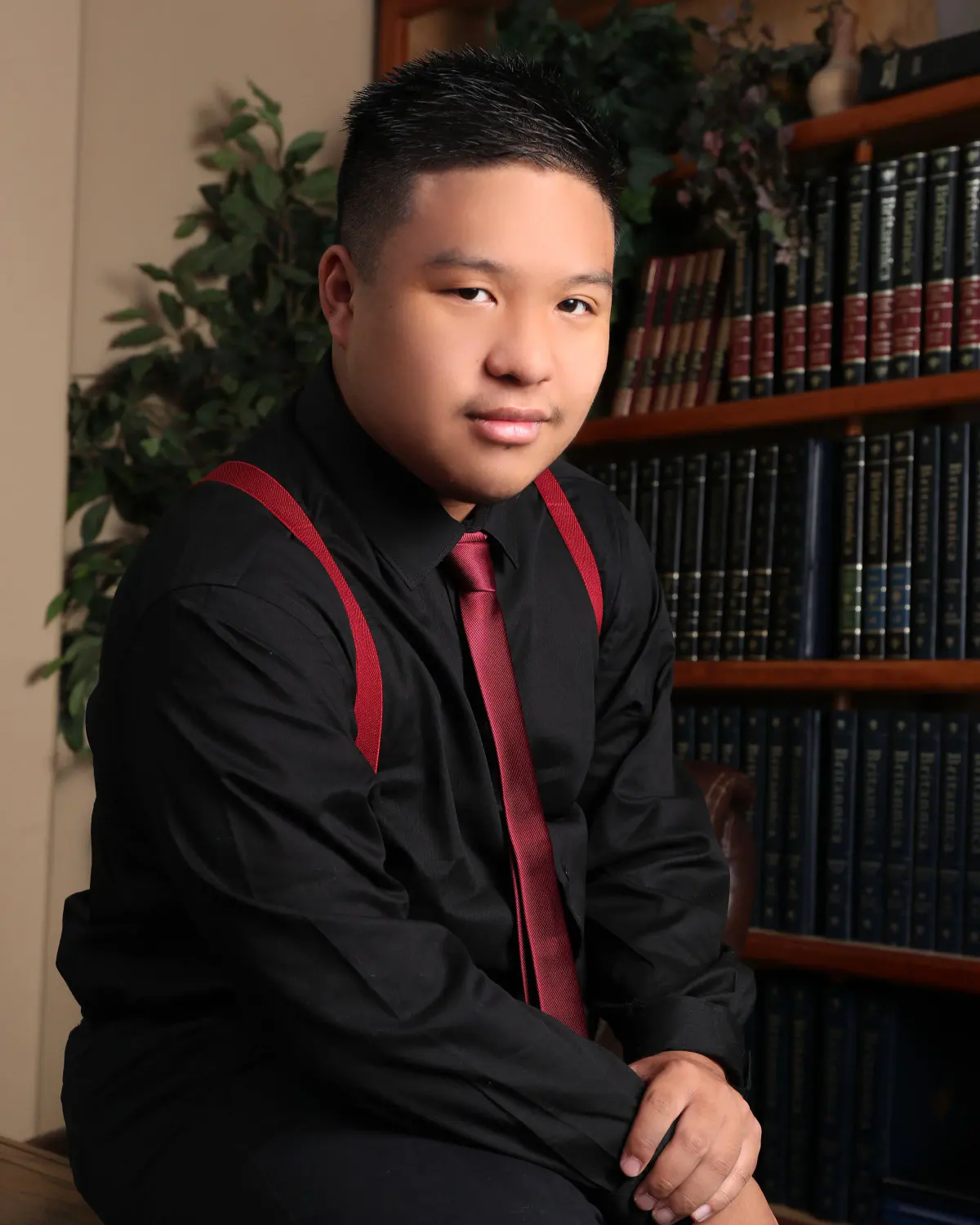 Senior young man in black shirt with red suspenders and red tie, seated, leaning forward in the library set with bookshelves and a potted plant