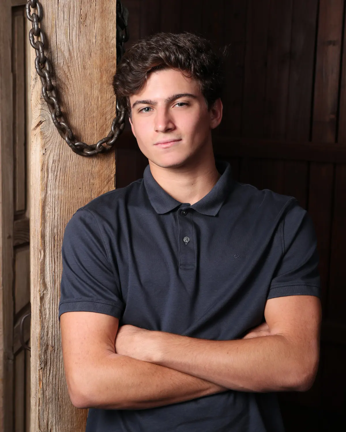 Senior young man with dark curly hair, navy polo, arms crossed, leaning against a weathered wooden post with a hanging iron chain