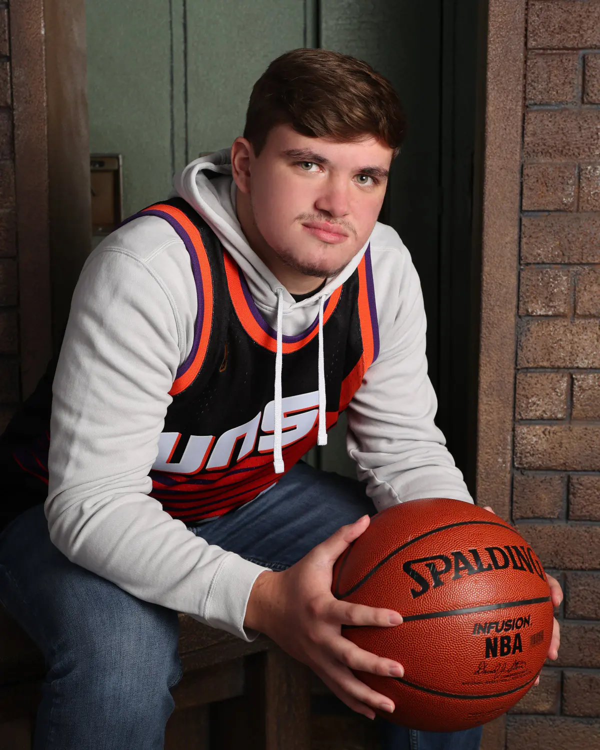 Senior young man in a gray hoodie under a purple Phoenix Suns-style jersey, seated, holding a Spalding basketball