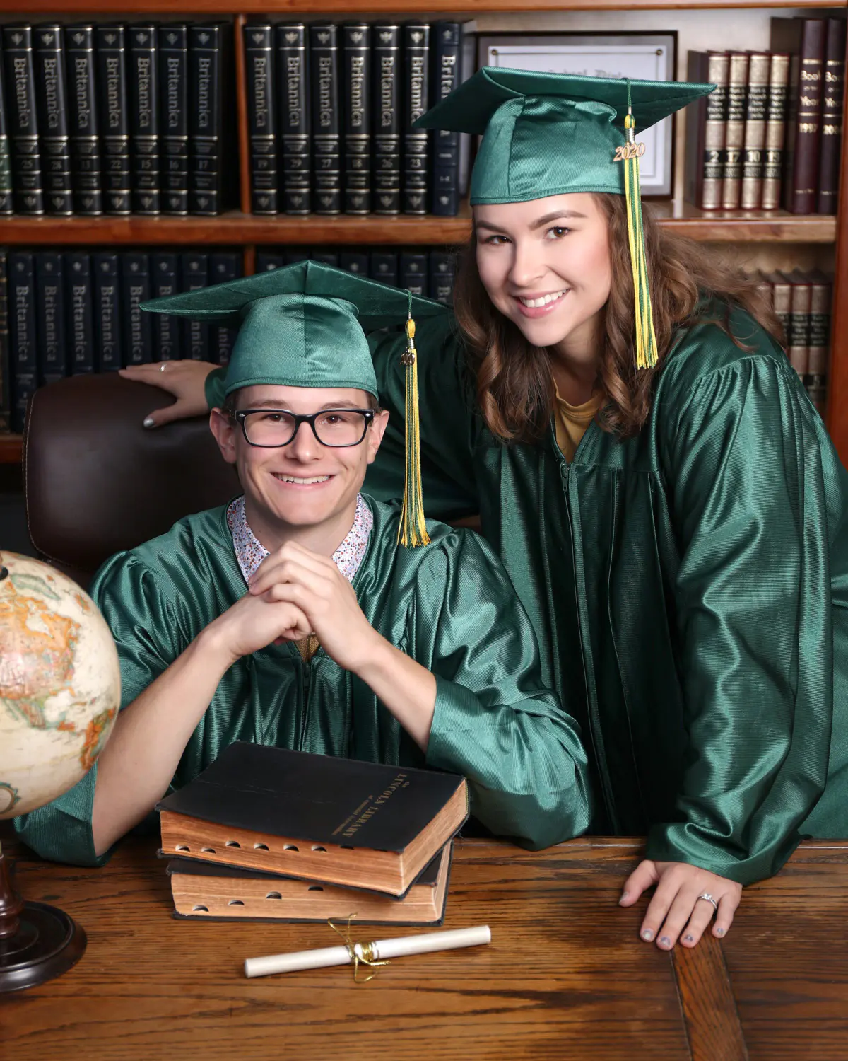 Two graduates in emerald green caps and gowns at a wooden desk in the library set, young man with glasses seated