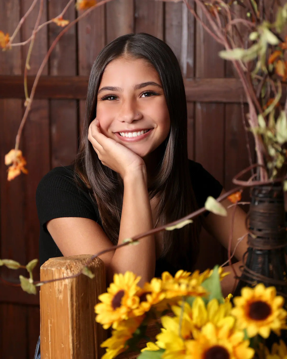 Smiling senior girl with long dark hair, black tee, chin resting on hand on a rustic wood fence post