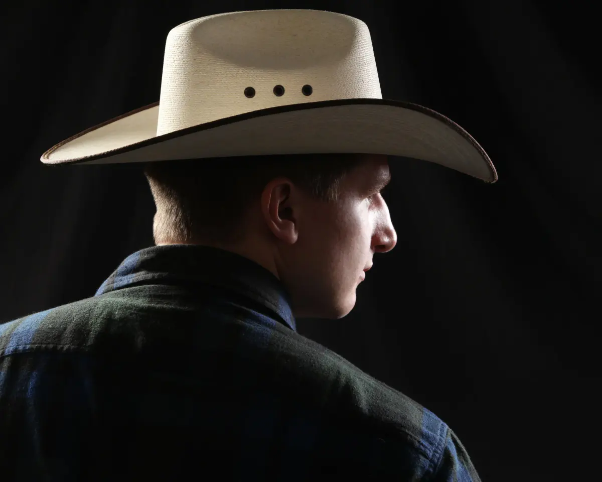 Over-the-shoulder profile of a senior young man in a white straw cowboy hat and dark plaid flannel, face turned in profi