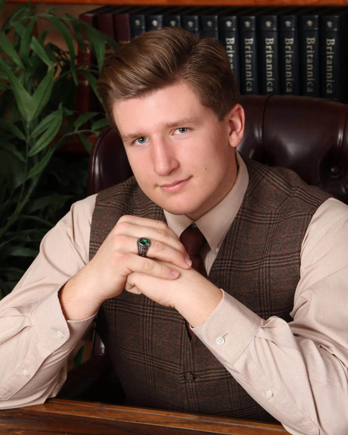 Senior young man in a brown tweed vest, pink shirt and tie with a class ring, hands clasped, seated at a desk on a library set with leather chair