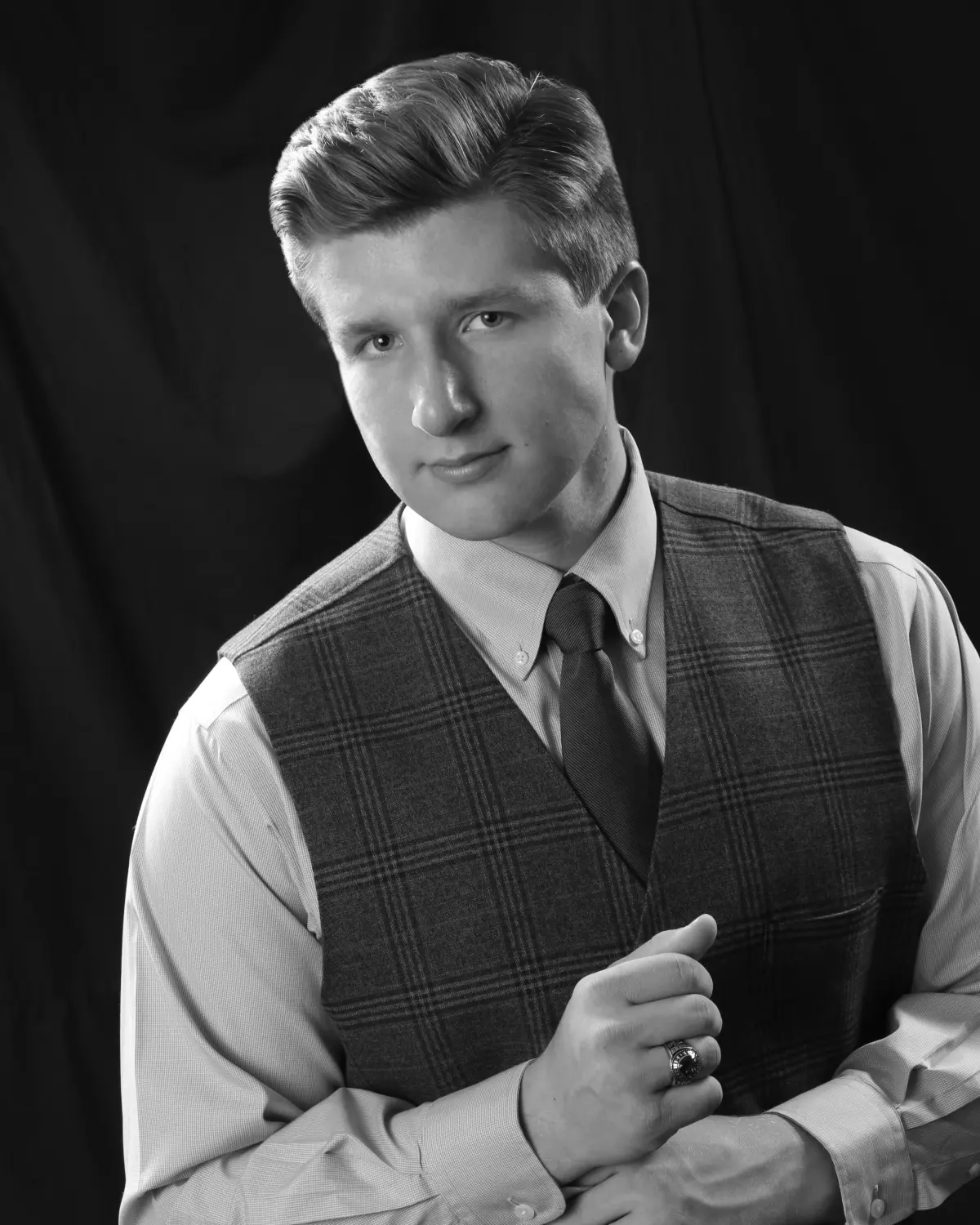 Black-and-white three-quarter portrait of a senior young man with swept pompadour in a plaid vest, button-down and tie, ringed hand at chest