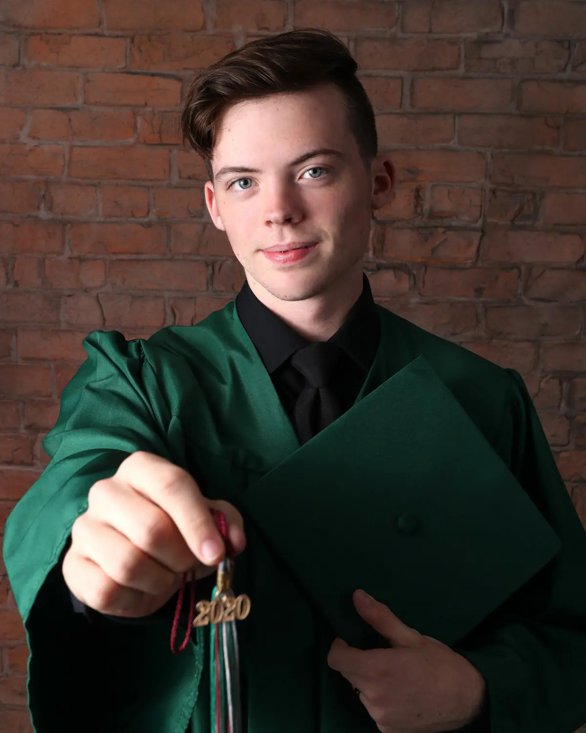 Senior young man in a forest-green graduation gown and black shirt holding his cap and a '2020' tassel toward camera, red brick backdrop