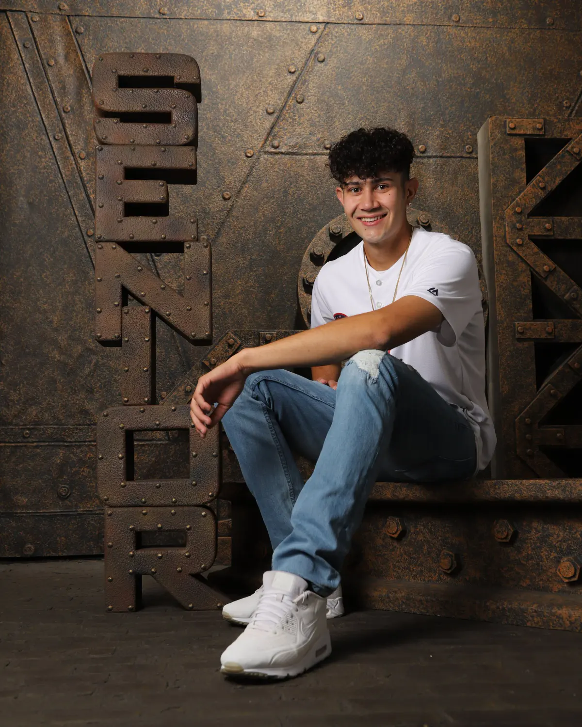 A senior seated on a rusted-metal studio set with tall SENIOR cut-out letters
