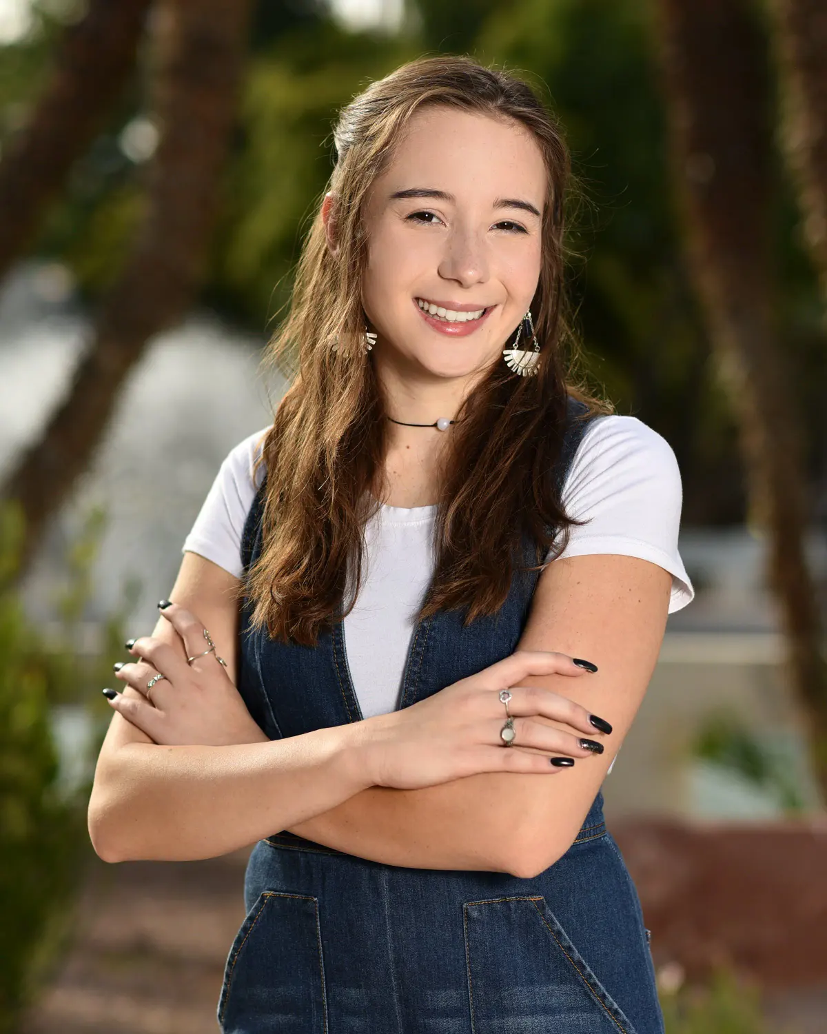 Smiling senior young woman with long brown hair in a white tee under denim overalls, arms crossed, outdoors with palm fronds