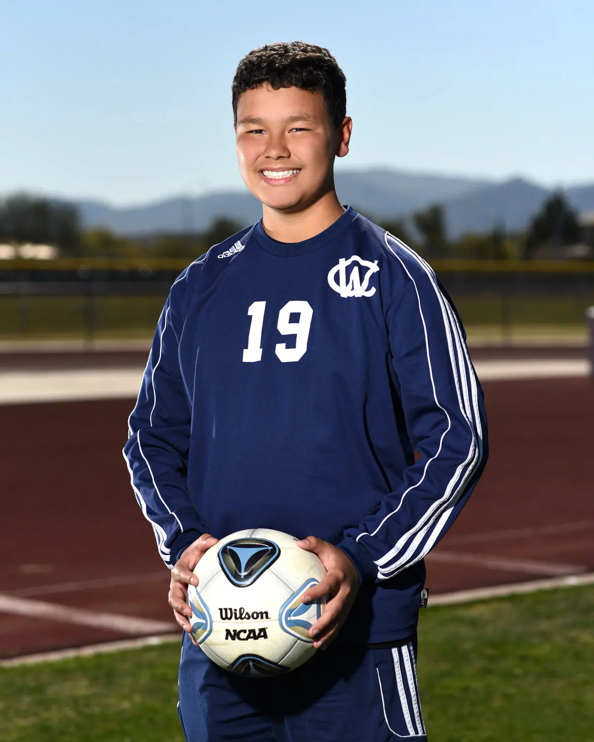 Teen boy in a navy long-sleeve soccer jersey, number 19 with 'WC' crest, holding a Wilson NCAA soccer ball, smiling