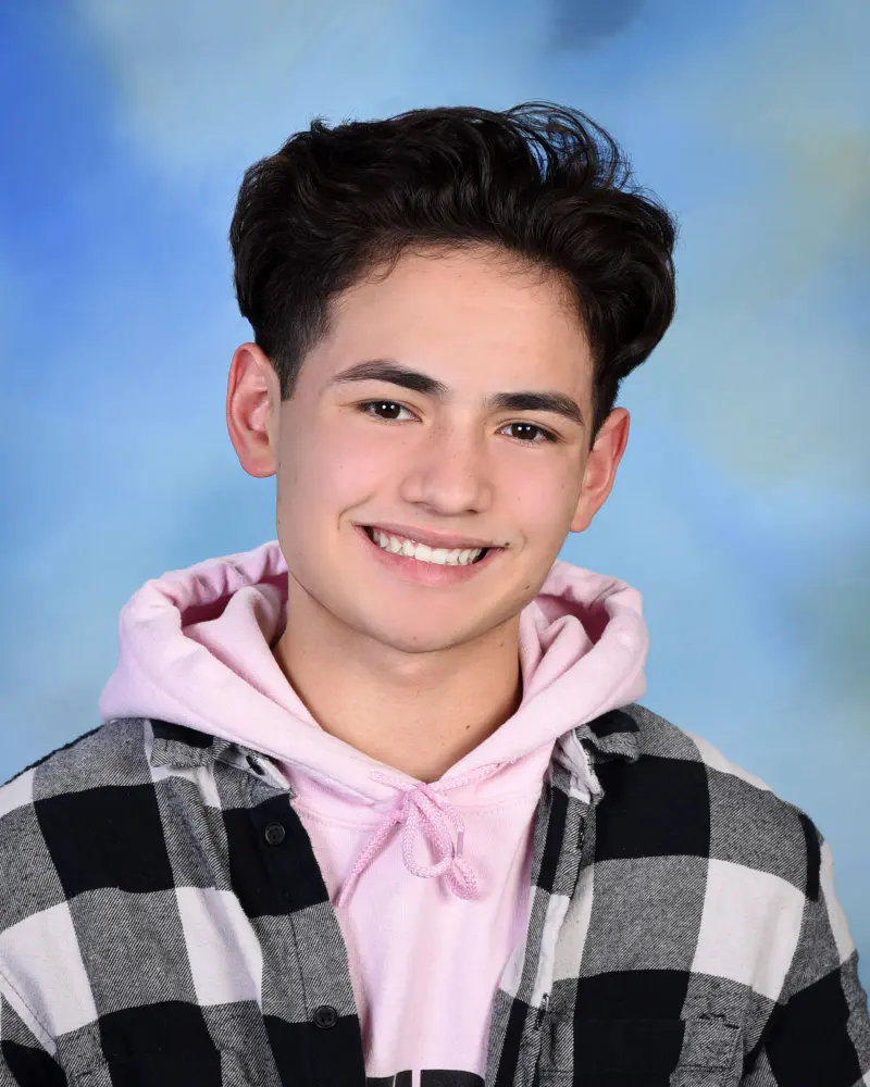 Smiling teen boy with dark wavy hair, pink hoodie under a black and white plaid flannel, head-and-shoulders on a mottled