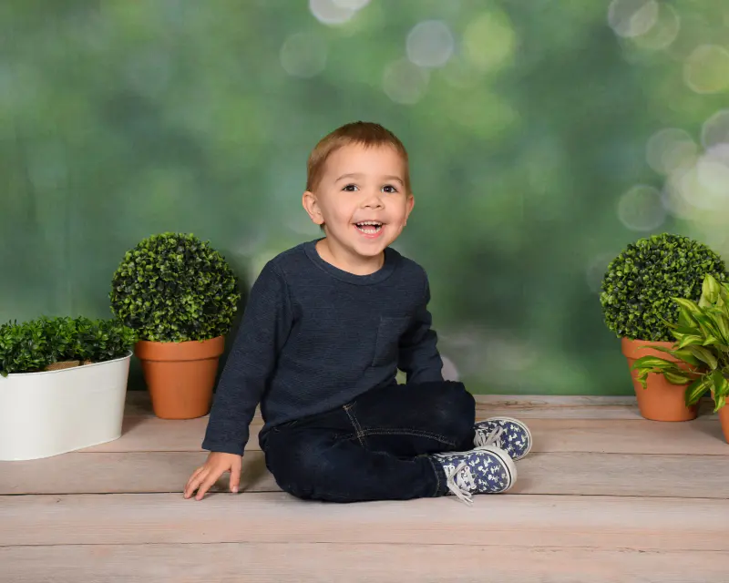 Laughing toddler/preschool boy in navy sweater and jeans sitting on whitewashed wood planks between potted topiary plant