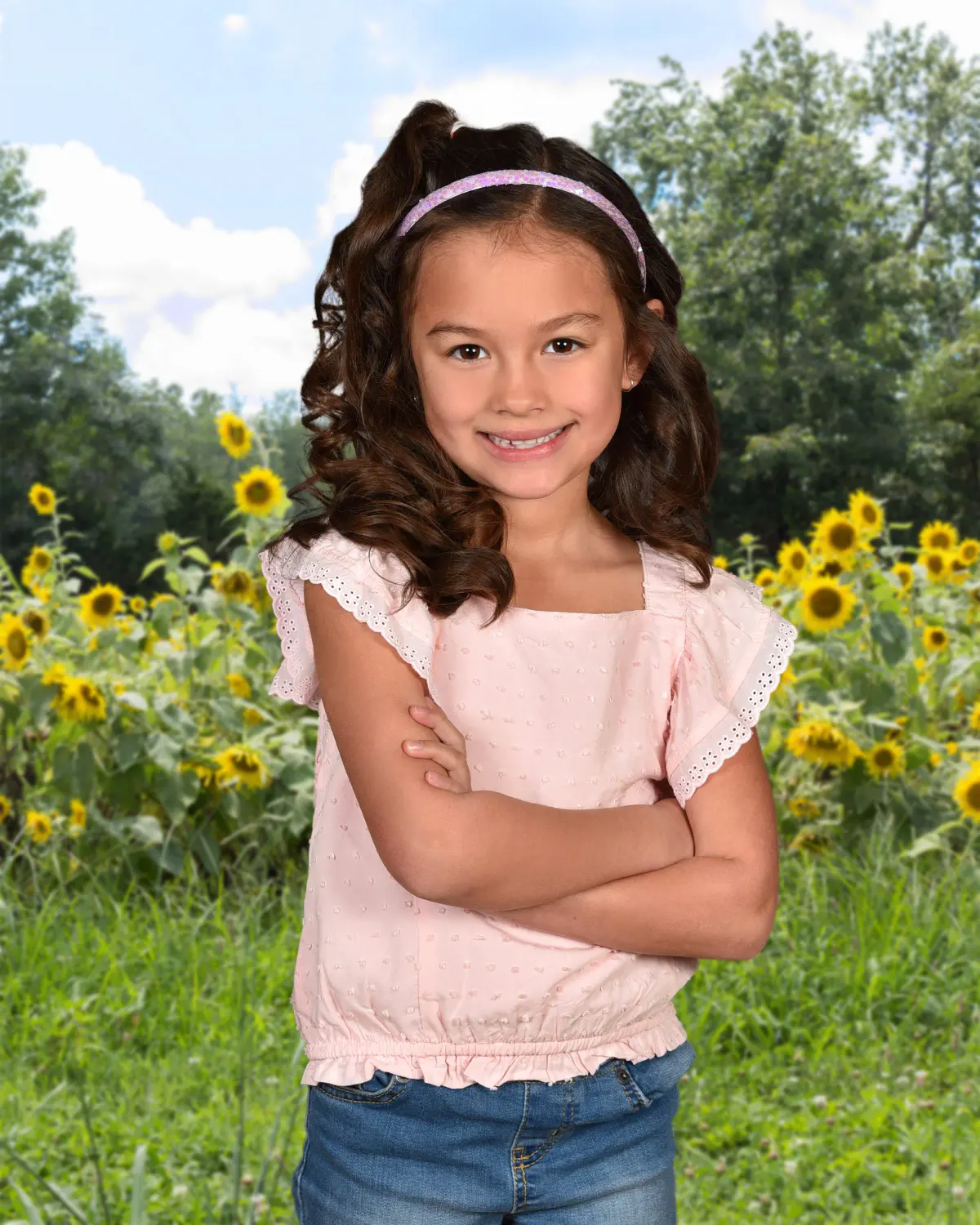 Young girl in a pink top and jeans with a headband, arms crossed, smiling, in front of a sunflower field and trees background