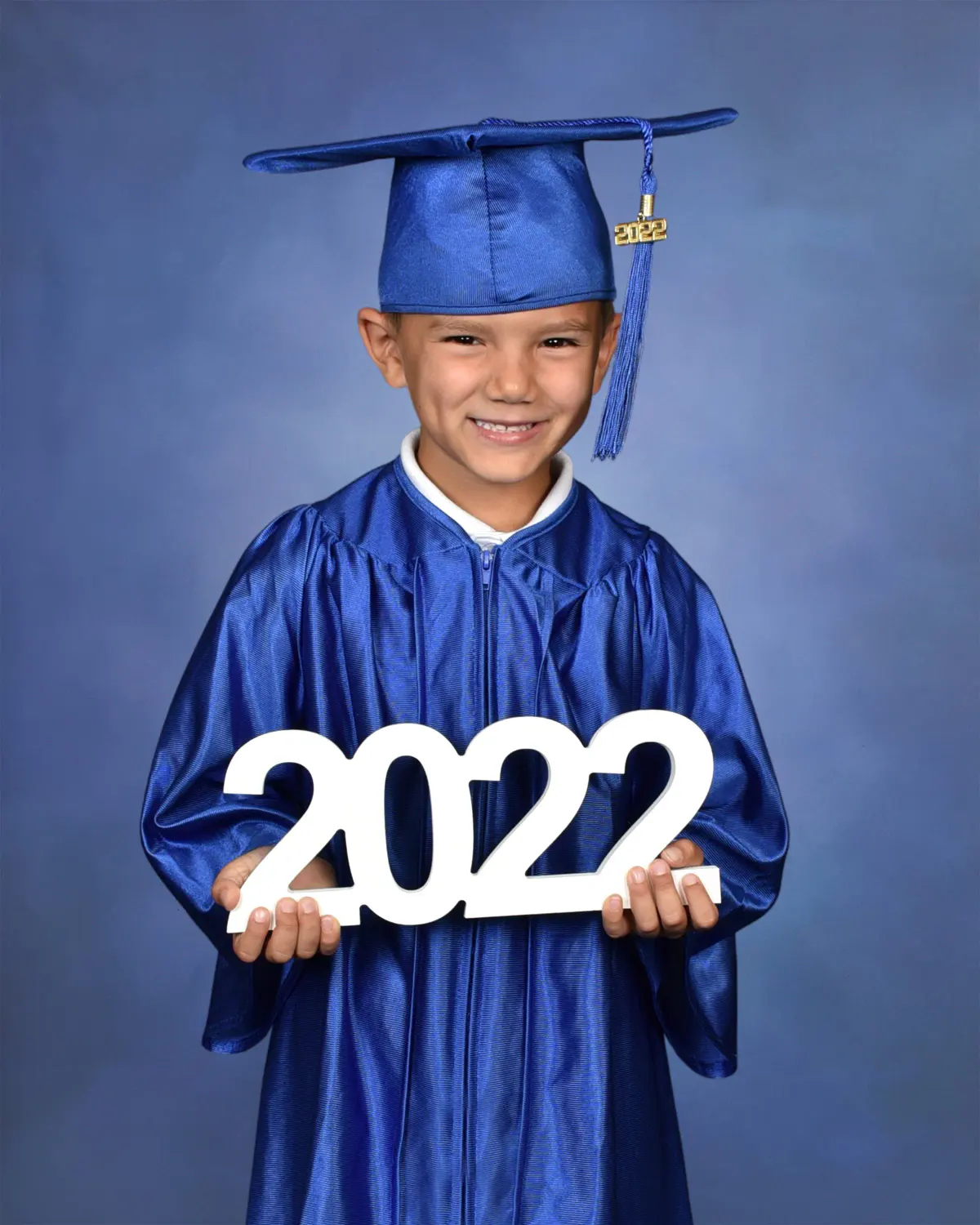 Young child in a shiny royal-blue cap and gown holding white '2022' numerals, on a blue mottled backdrop