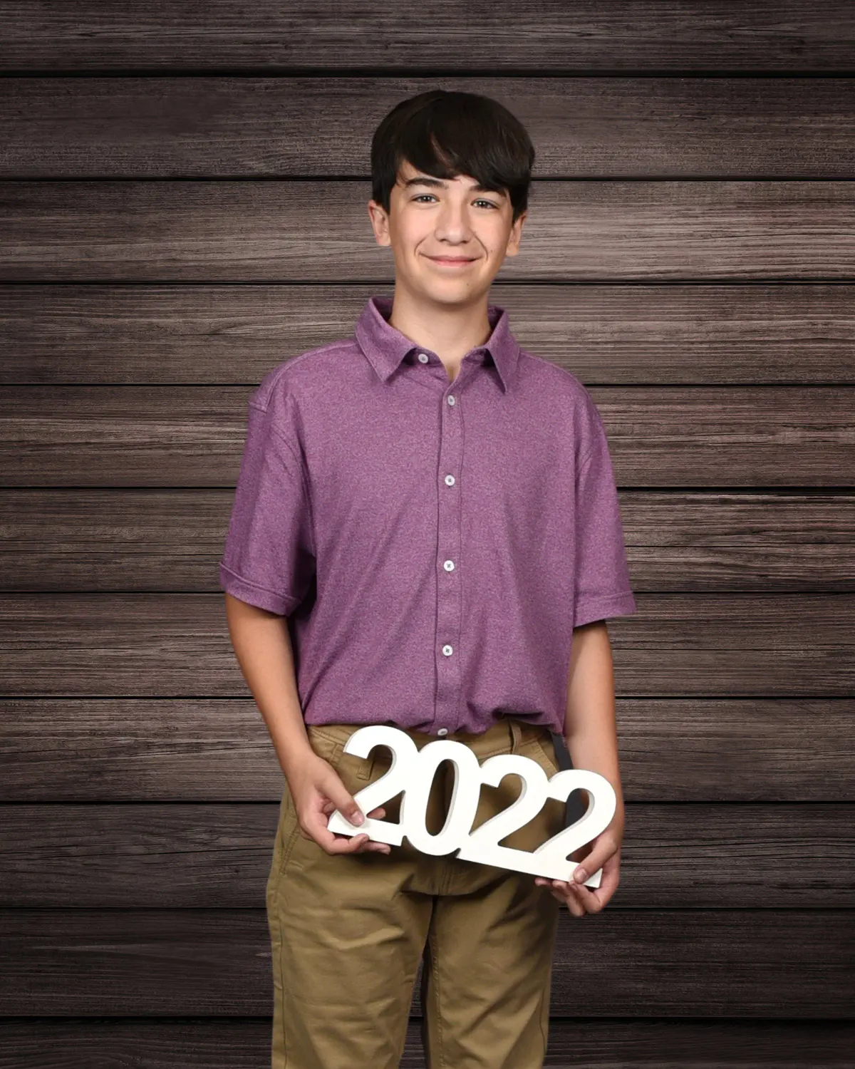 Preteen boy in a purple polo and khakis holding white '2022' numerals, on a dark wood-plank backdrop