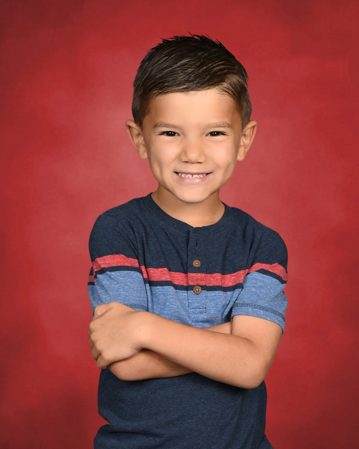 Young boy with arms crossed in a navy striped henley tee, big smile, on a saturated red mottled backdrop