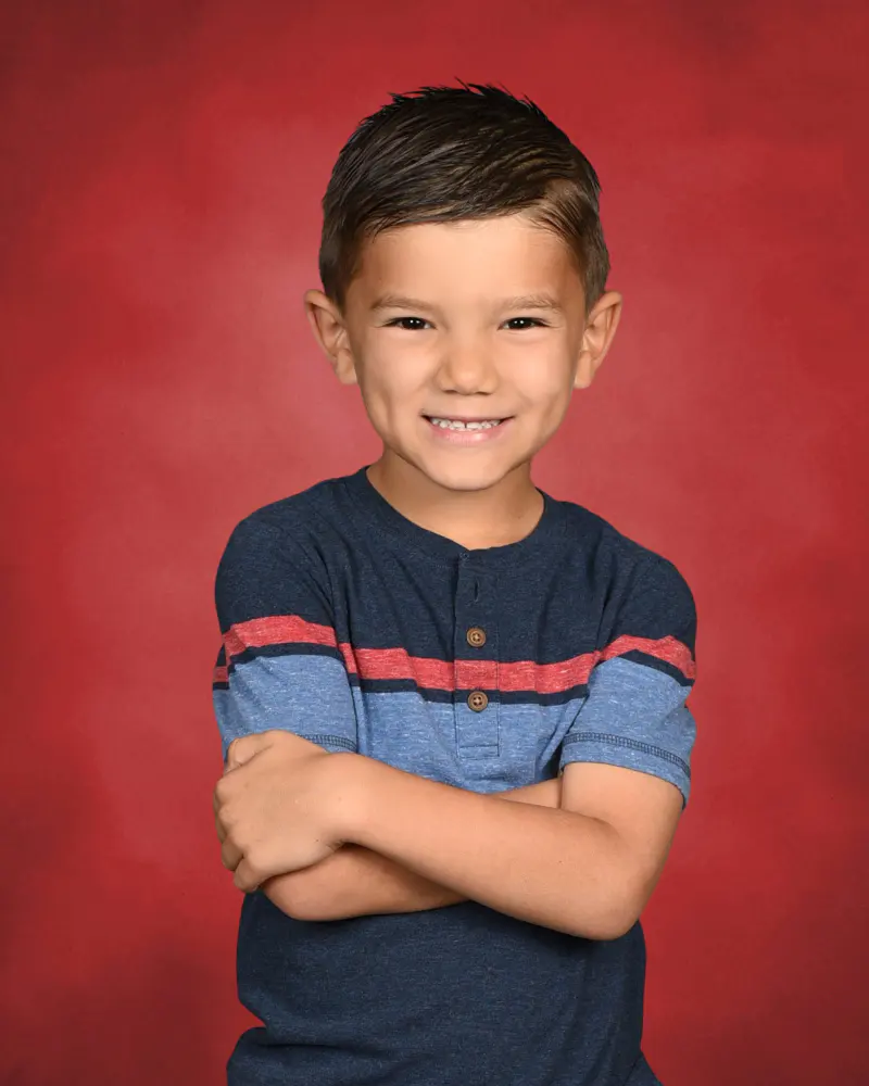 School portrait of a smiling boy in a striped tee on a red backdrop