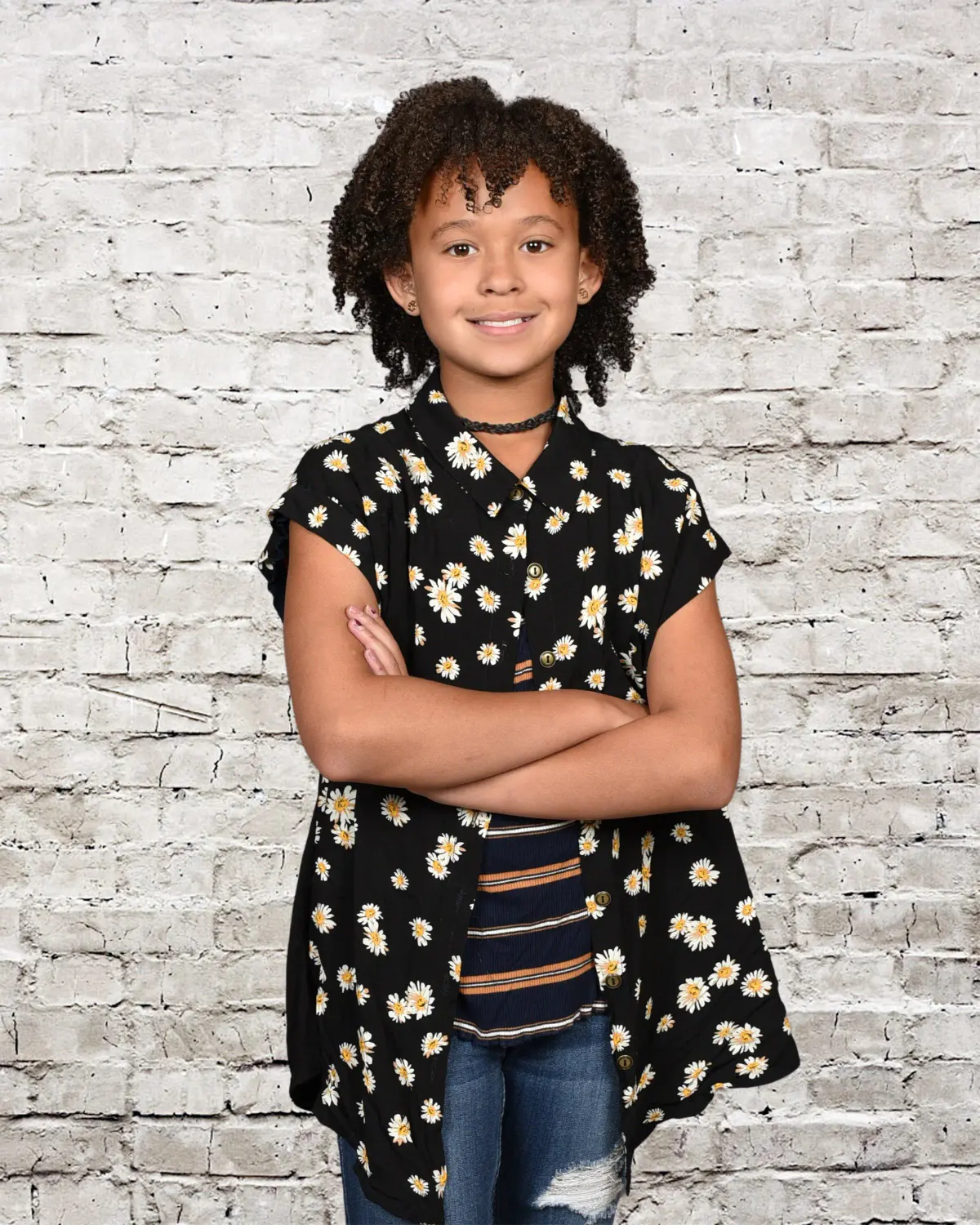 Girl with curly hair in a black daisy-print top over a striped shirt and ripped jeans, arms crossed, on a white-painted brick backdrop