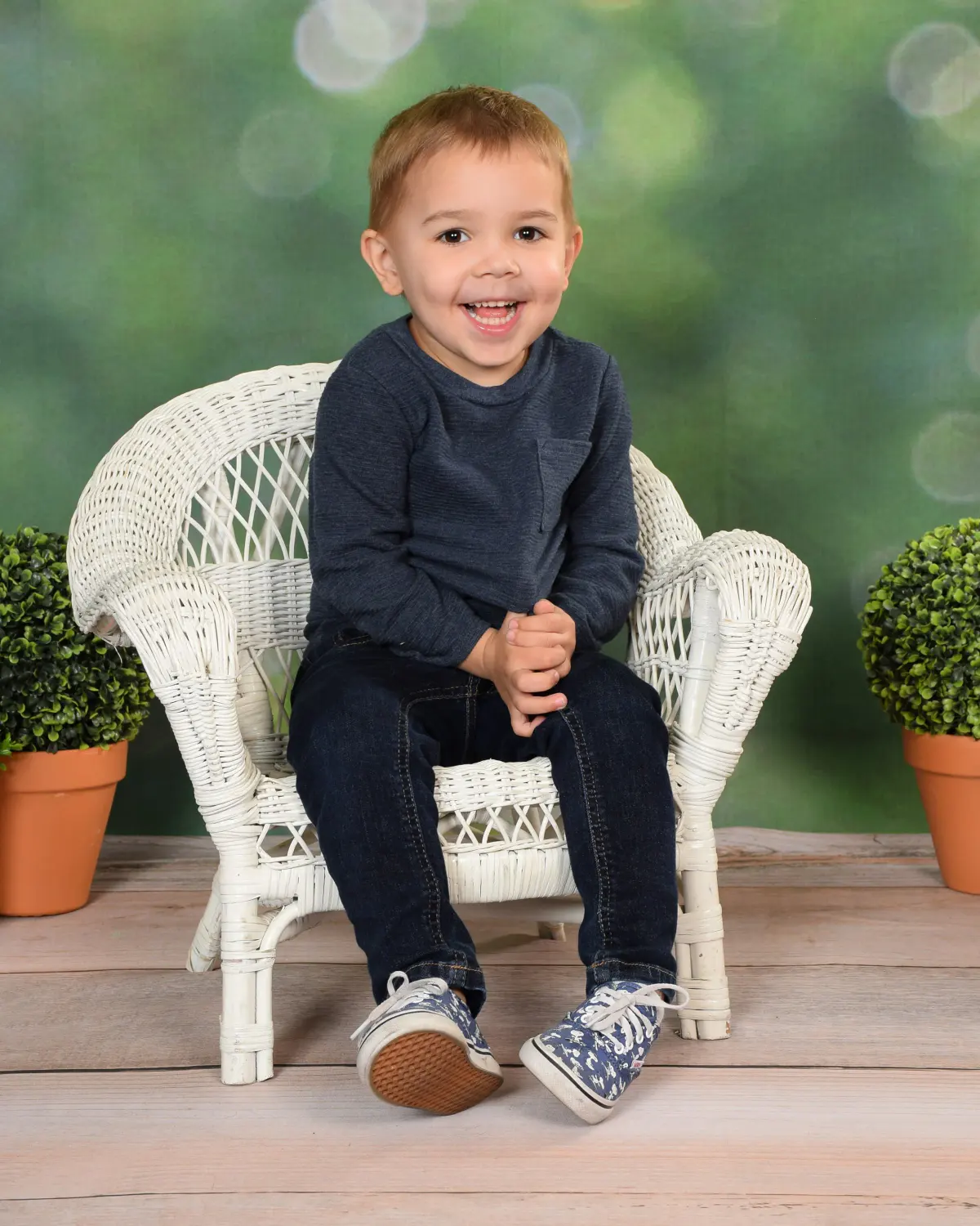 Toddler-age child in navy sweater and jeans laughing on a small white wicker chair, flanked by potted topiaries