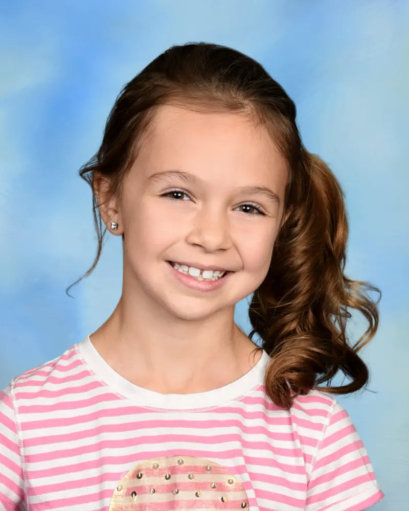 School portrait of a smiling girl in a pink striped tee on a blue backdrop