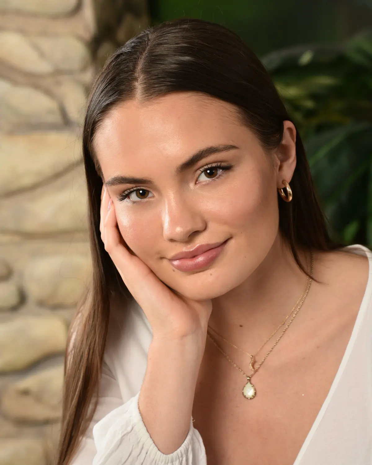 Senior girl with long dark hair, white blouse and layered gold necklaces, hand resting on cheek, soft closed-mouth smile