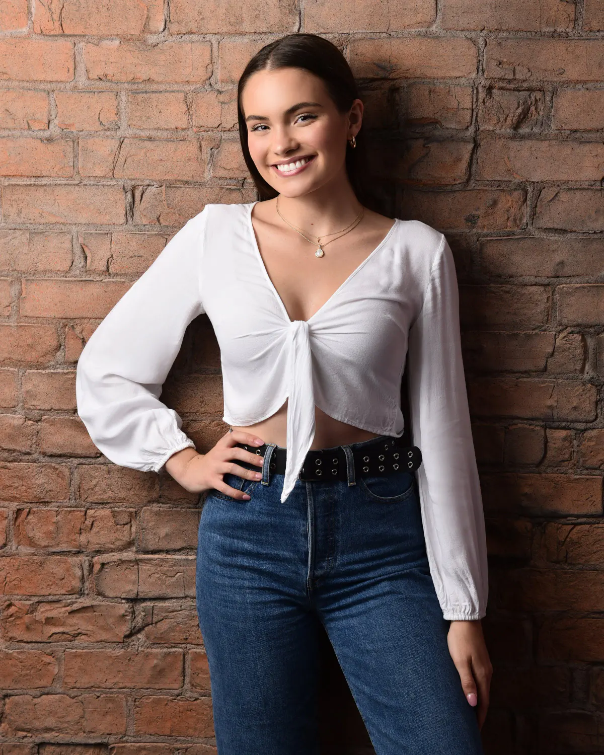 Senior girl, hand on hip, white tie-front blouse and jeans, against a red-brown brick wall