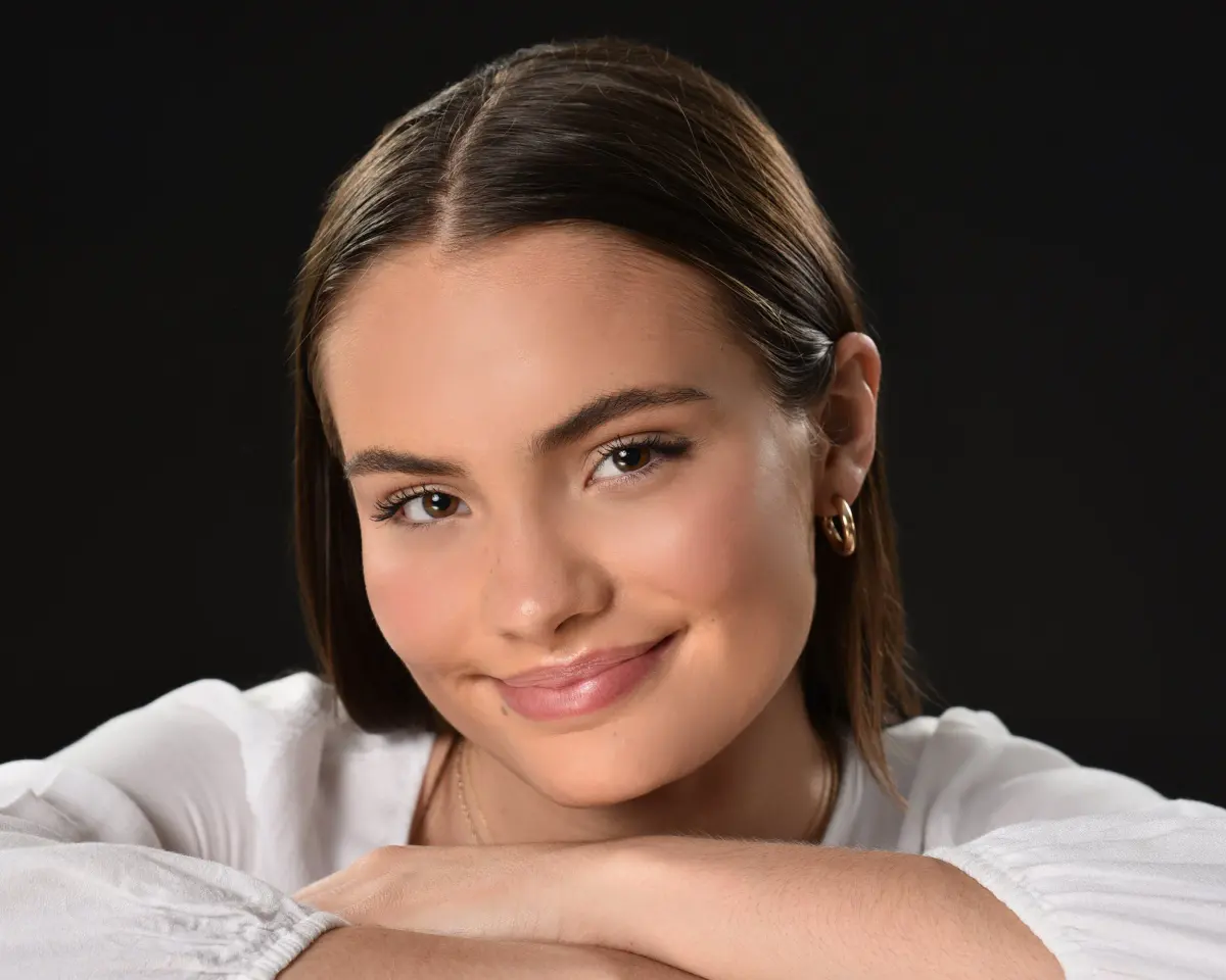 Same senior girl, tight headshot, chin resting on folded arms in a white blouse, head tilted, soft smile, gold hoop earr