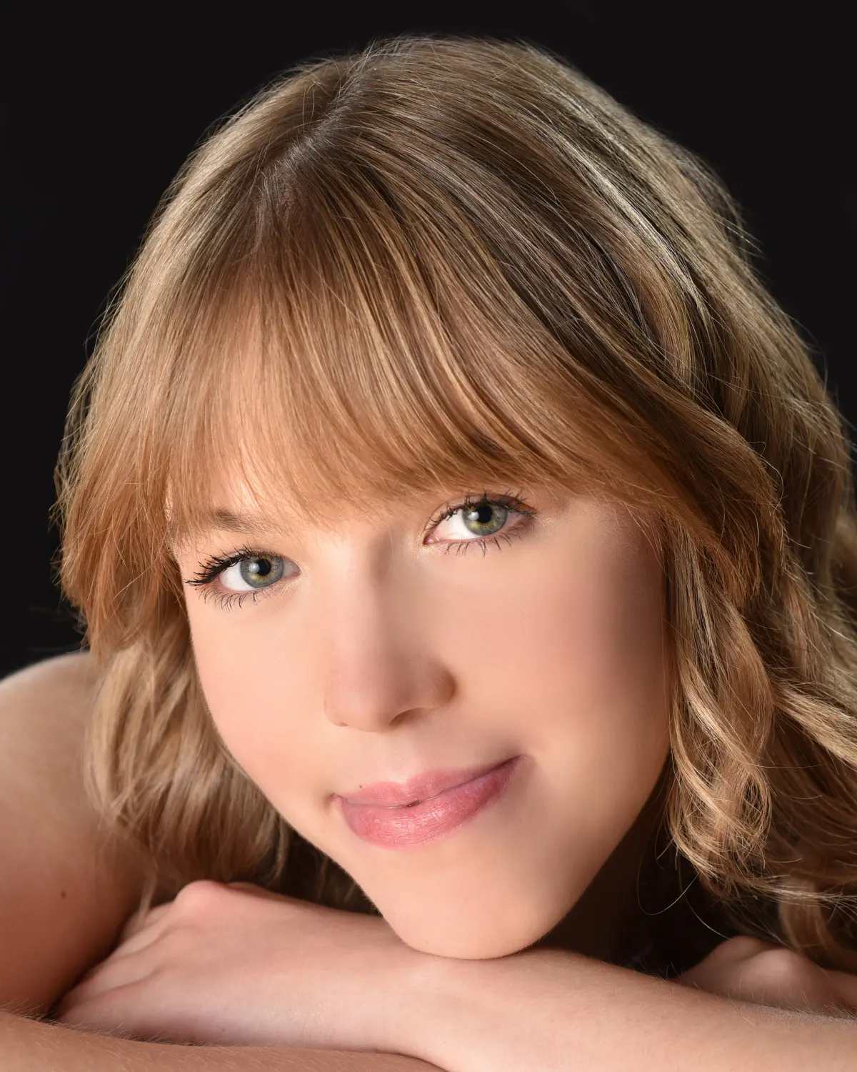 Tight close-up beauty portrait of a young woman with honey-blonde hair and bangs, chin resting on folded arms, bare shou