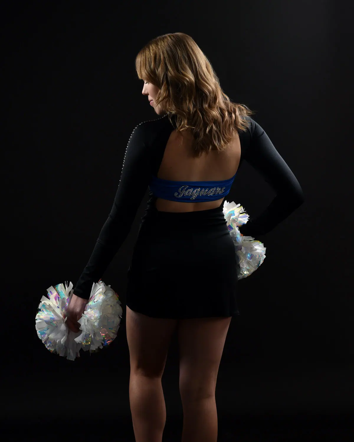 Young woman shot from behind, looking over her shoulder, wearing black open-back pom uniform with blue rhinestone 'Jaguars' band